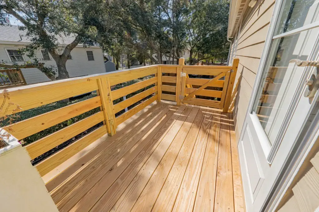 elevated-wooden-deck-and-railing-with-a-gate-on-the-mocha-house-beside-the-white-trim-door-with-big-trees-and-houses-in-the-background-in-outer-banks-nc