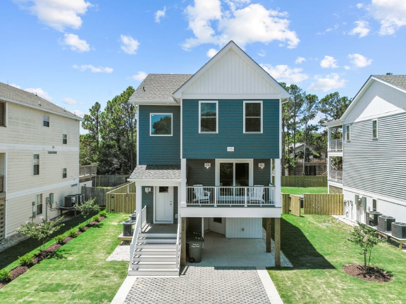 front-view-of-the-residential-house-with-siding-and-under-deck-with-white-white-railing-and-outdoor-white-two-chairs-featuring-houses-and-blue-sky-with-clouds-view-in-outer-banks-nc