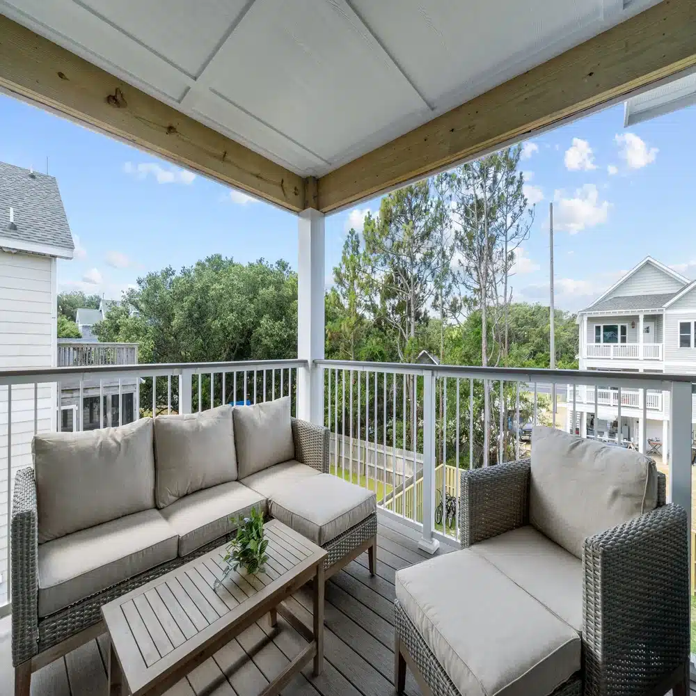 outdoor-living-area-with-couch-and-table-on-the-elevated-deck-with-white-railings-featuring-houses-and-trees-in-the-background-in-outer-banks-nc